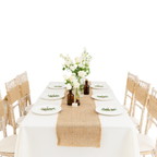 Dining table set with white tablecloth, burlap runner, plates, and flowers on a white background